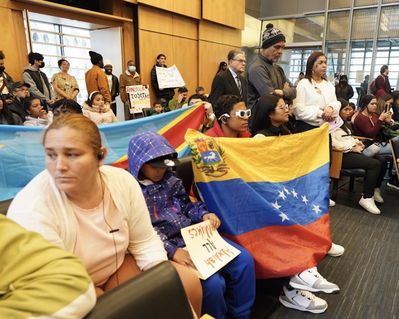 caption: Adriana Figueira (standing in white to the right) is one of the community leaders for the group of asylum-seekers, primarily Venezuelans who have Temporary Protected Status. That means they have special rights as immigrants to get work authorization. She spoke at Seattle City Hall on Jan. 30, 2024.