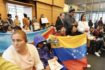 caption: Adriana Figueira (standing in white to the right) is one of the community leaders for the group of asylum-seekers, primarily Venezuelans who have Temporary Protected Status. That means they have special rights as immigrants to get work authorization. She spoke at Seattle City Hall on Jan. 30, 2024.