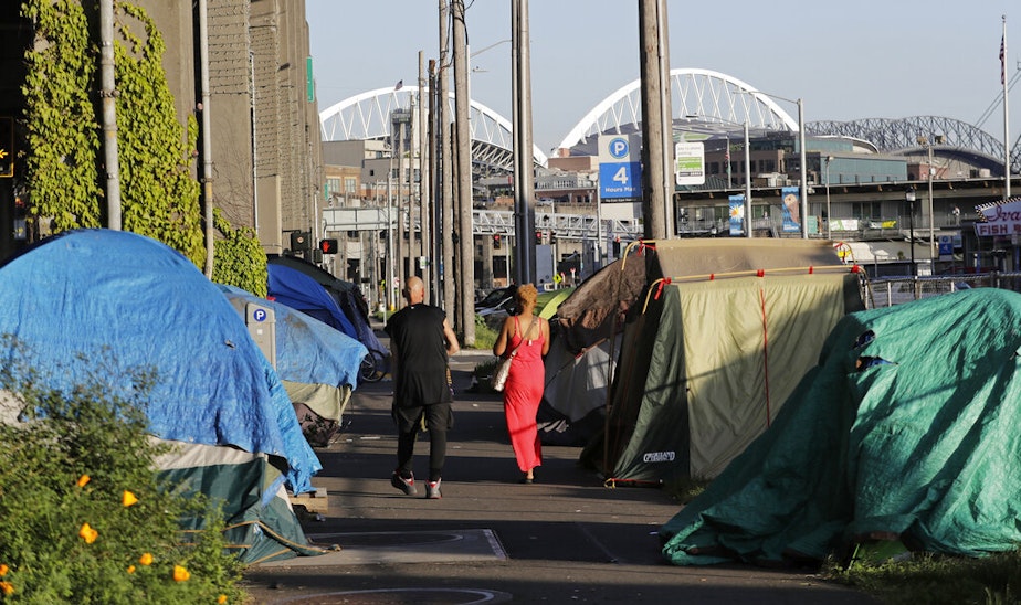 caption: In this May 7, 2018 file photo, tents used by homeless people are shown on either side of a sidewalk in Seattle with CenturyLink and Safeco fields in the background. 