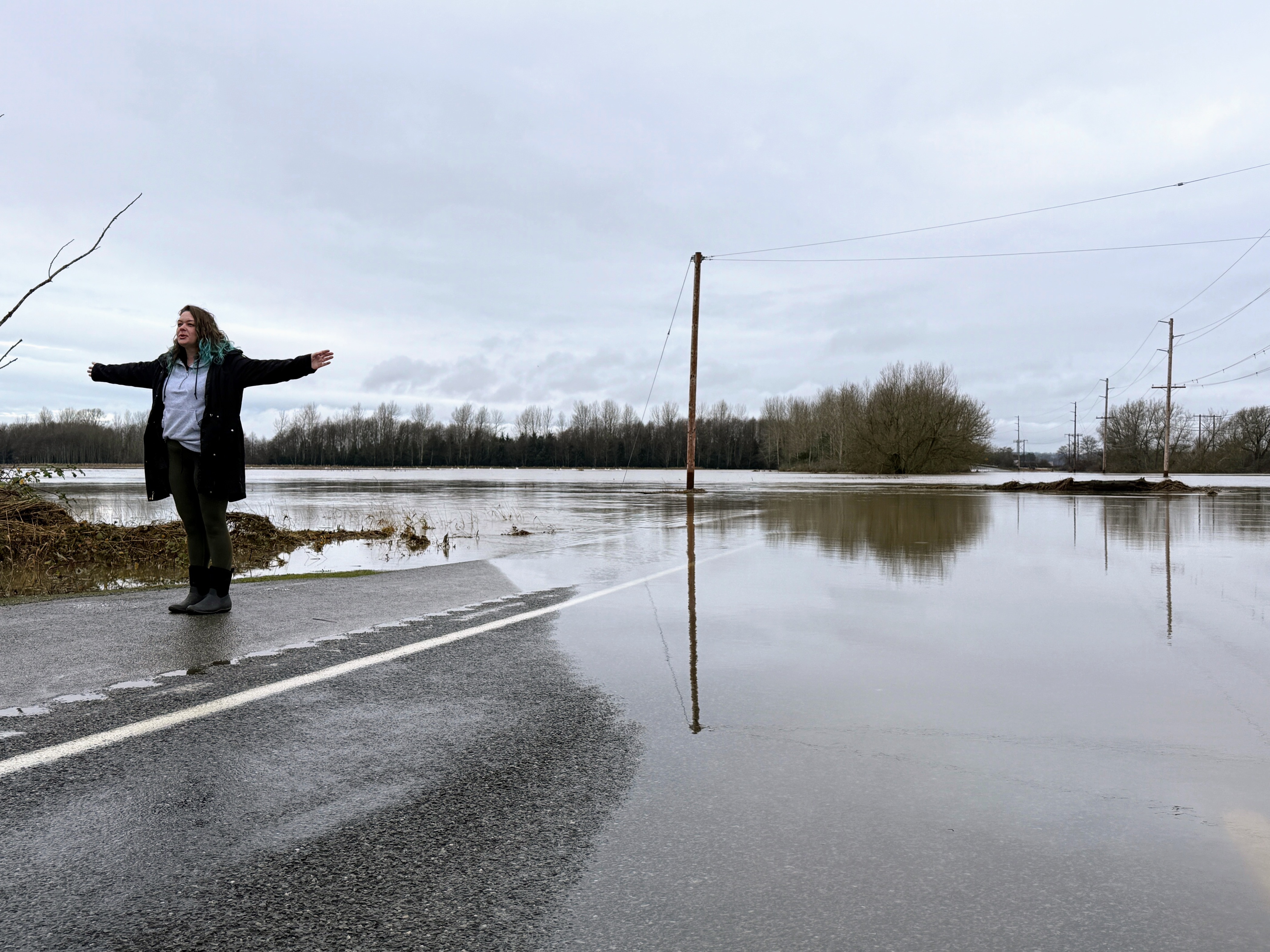 caption: Maddie Crerar of Bellingham stands on Slater Road next to the swollen Nooksack River near Ferndale, Washington, on Dec. 12, 2025.