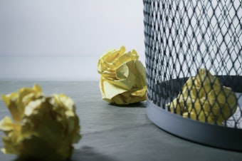 caption: Crumpled papers sit next to an office trash can.