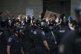 caption: Protesters are shown raising their hands in the air while chanting, 'hands up, don't shoot,' toward Seattle police officers on Saturday, May 30, 2020, near the intersection of 5th and Pine Streets in Seattle. Thousands gathered in a protest following the violent police killing of George Floyd, a Black man who was killed by a white police officer who held his knee on Floyd's neck for 8 minutes and 46 seconds, as he repeatedly said, 'I can't breathe,' in Minneapolis on Memorial Day. 