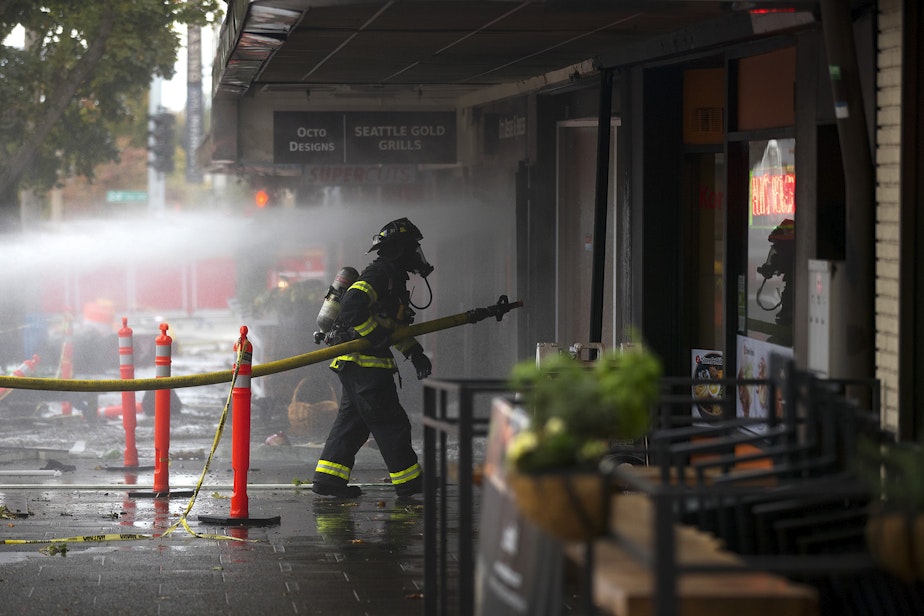 caption: Firefighters work to put out a fire on Monday, October 7, 2019, at the intersection of NW Market Street and 24th Avenue Northwest in Seattle.