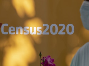 caption: A person wearing a mask walks past a sign encouraging people to complete the 2020 census in Los Angeles.