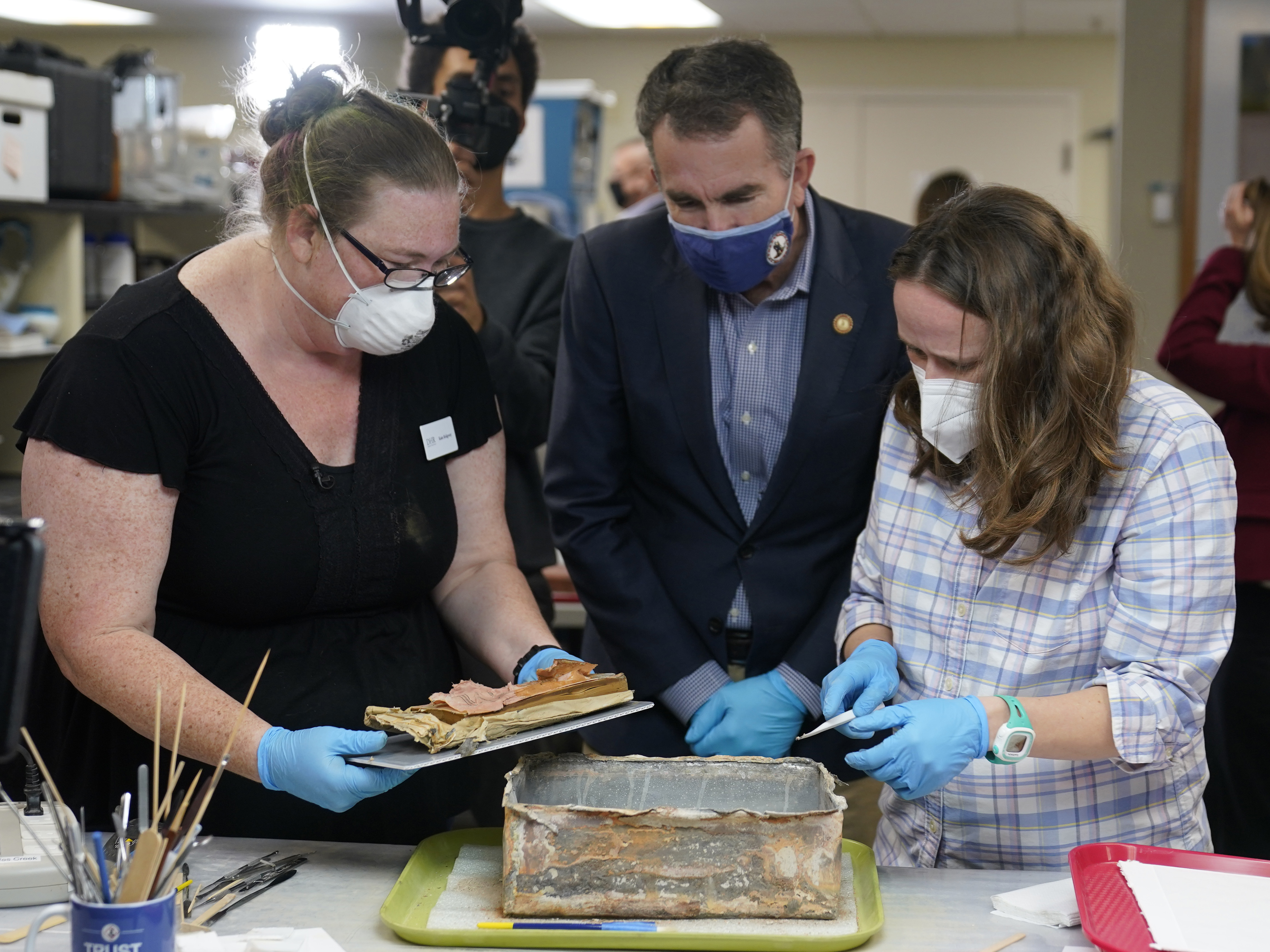 caption: Virignia Gov. Ralph Northam ( center) watches as lead conservator for the Virginia Department of Historic Resources, Kate Ridgway (left) and Sue Donovon, conservator for Special Collections for the University of Virginia, remove the contents of a time capsule  from the pedestal that once held the statue of Confederate General Robert E. Lee.