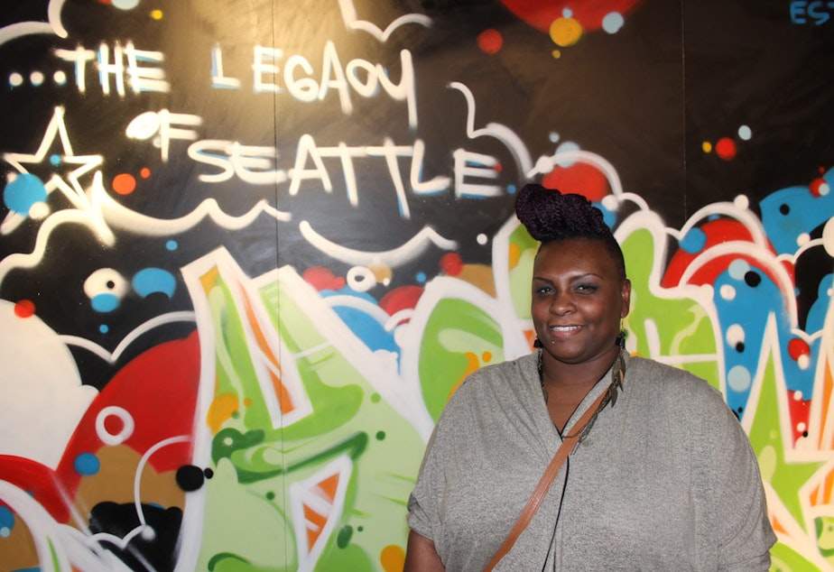 caption: Jazmyn Scott stands in front of a mural created for MOHAI by SPECSWIZARD who has been making art and beats in Seattle since 1978.