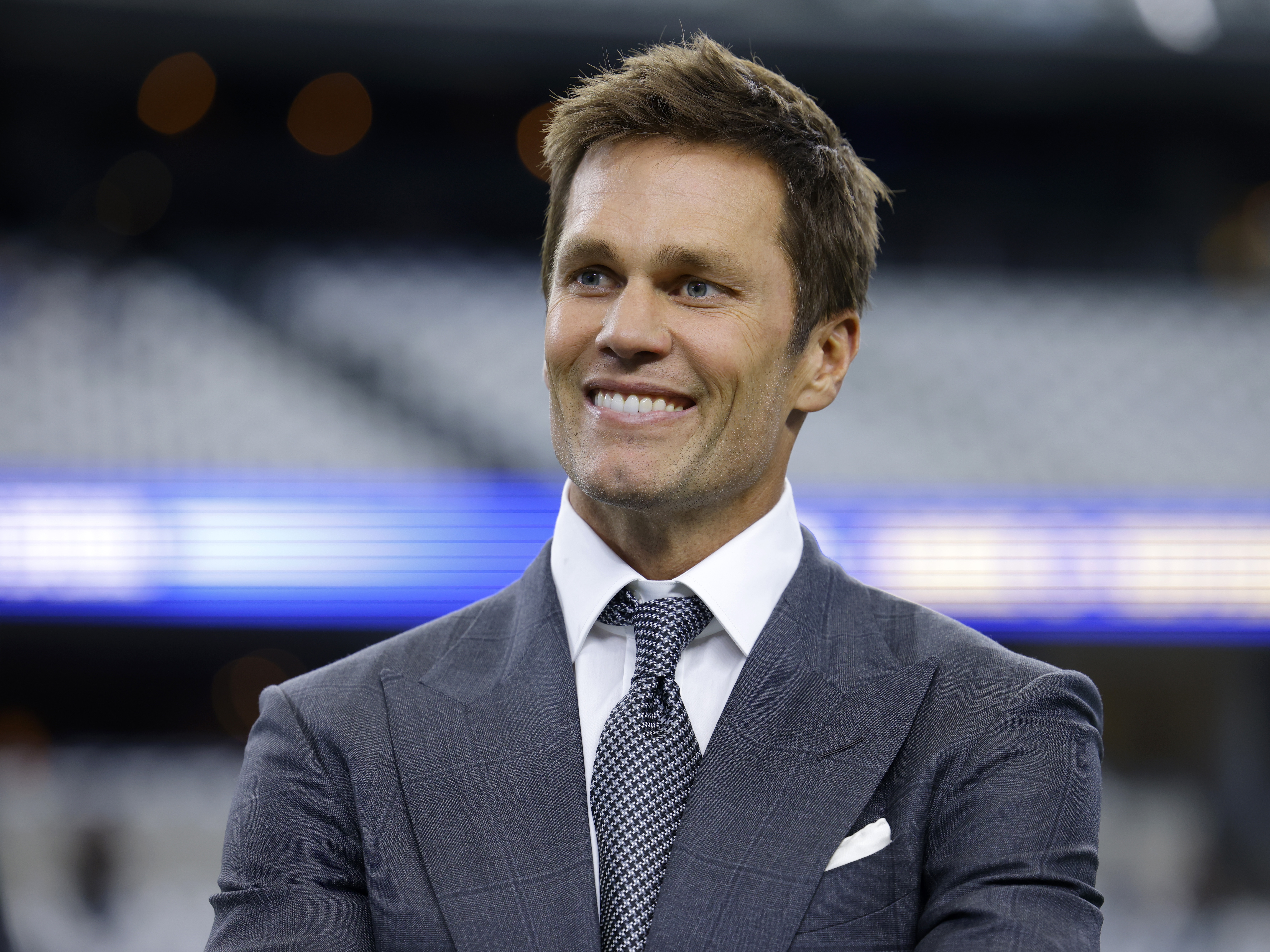 caption: Fox Sports lead NFL analyst Tom Brady stands on the field before a game between the Detroit Lions and the Dallas Cowboys in Arlington, Texas, on Sunday. The former NFL star quarterback has purchased a minority stake in the Las Vegas Raiders.