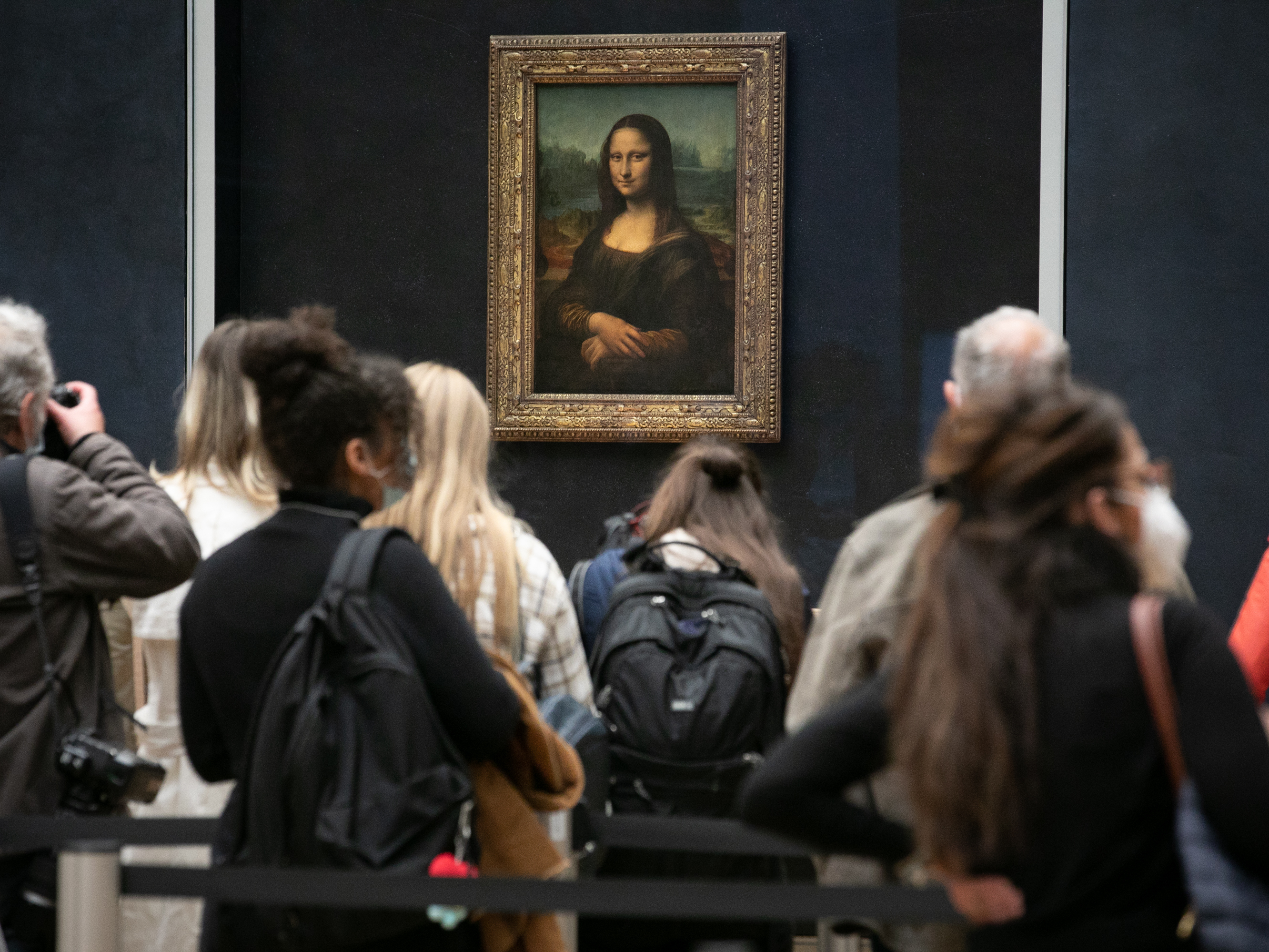 caption: Visitors observe the painting the <em>Mona Lisa</em> by Italian artist Leonardo da Vinci on display in a gallery at Louvre on May 19, 2021 in Paris, France.