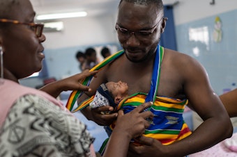 caption: New father Yappe Pako gets help with his kangaroo care carrier from a midwife. His newborn son is named Ambo Crisostome. They're in the kangaroo care ward at the University Hospital Medical Center at Treichville in the Ivory Coast. A new program teaches the technique to moms and dads. It's especially beneficial for preterm and low birthweight babies.