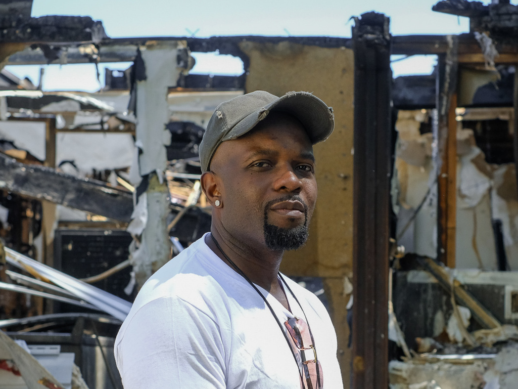 caption: Trevon Ellis, owner of Fade Factory, stands in front of the building where his barbershop was destroyed in Minneapolis. Sometime late Saturday, someone broke into the shop and lit the place on fire.