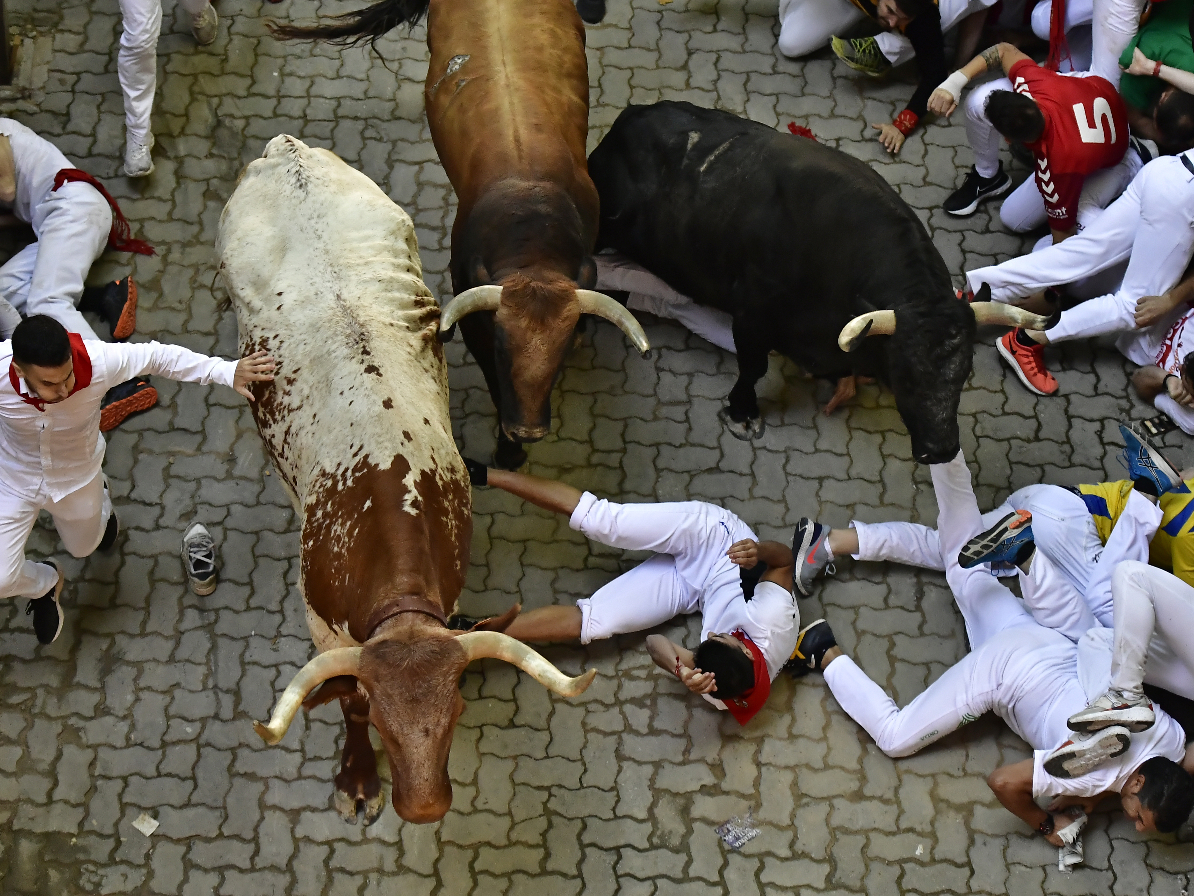 caption: Runners fall during the running of the bulls at the San Fermin Festival in Pamplona, northern Spain, Monday, July 11, 2022.