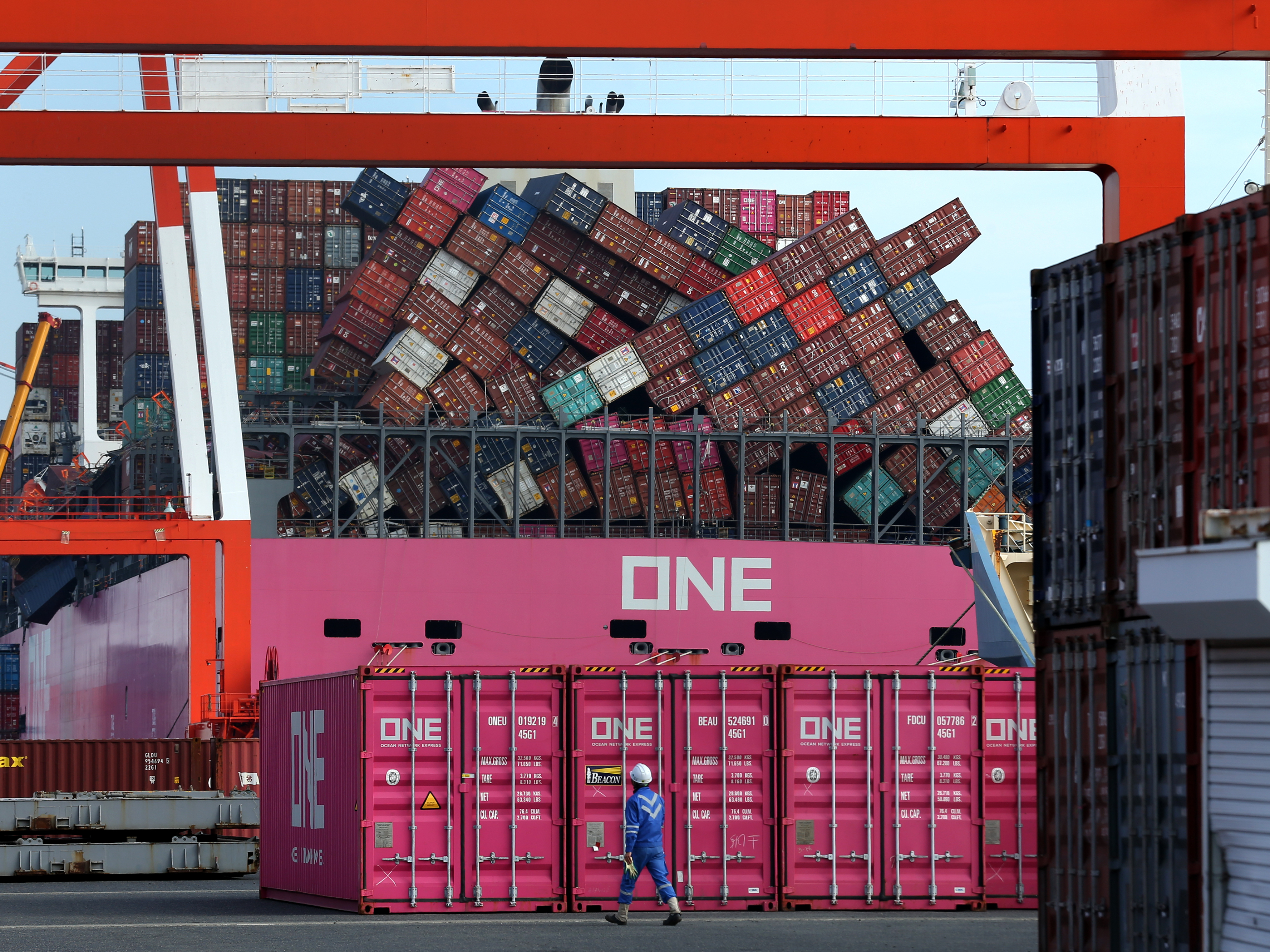 caption: A worker walks past the ONE Apus container ship and its dislodged containers at the Kobe Port in Japan in December. The vessel suffered a massive stack collapse and lost 1,816 containers at sea during severe weather on Nov. 30.
