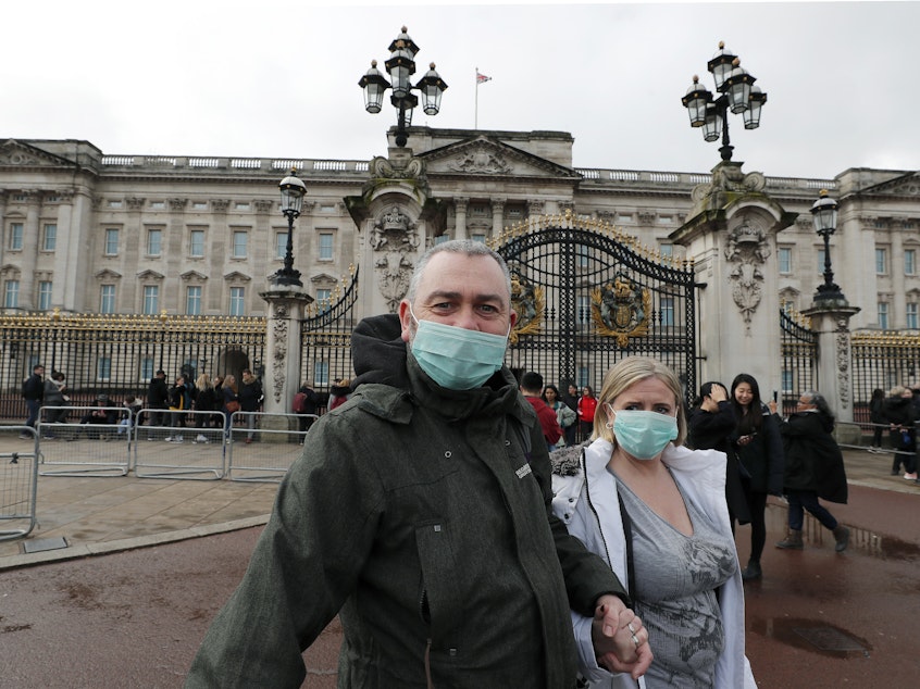 caption: A couple wear face masks as they visit Buckingham Palace in London on Saturday. After criticism for responding slowly, the United Kingdom has urged the public to avoid unnecessary contact.