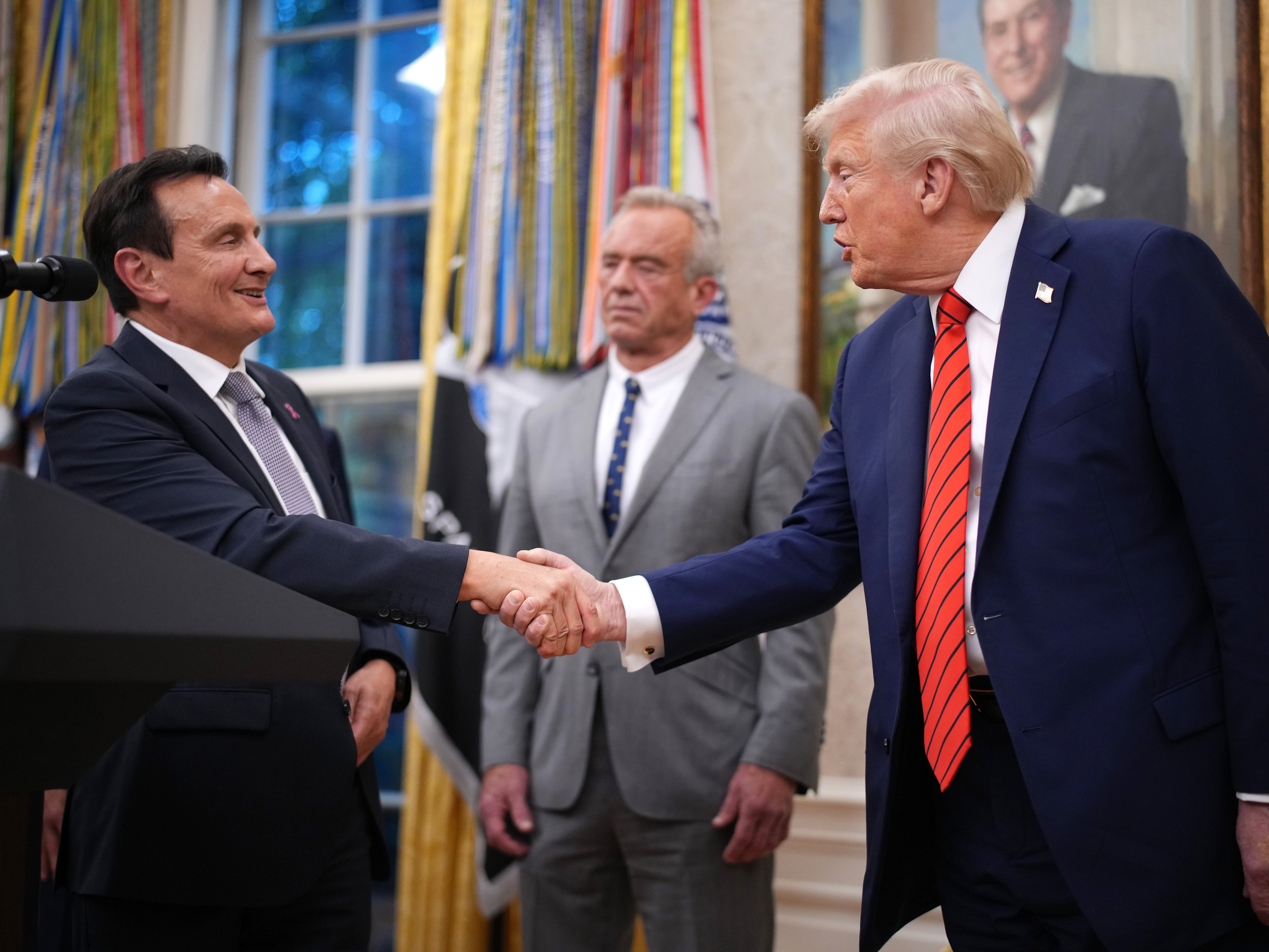 caption: AstraZeneca CEO Pascal Soriot shakes hands with President Trump in the Oval Office, as Health and Human Services Secretary Robert F. Kennedy Jr. looks on.