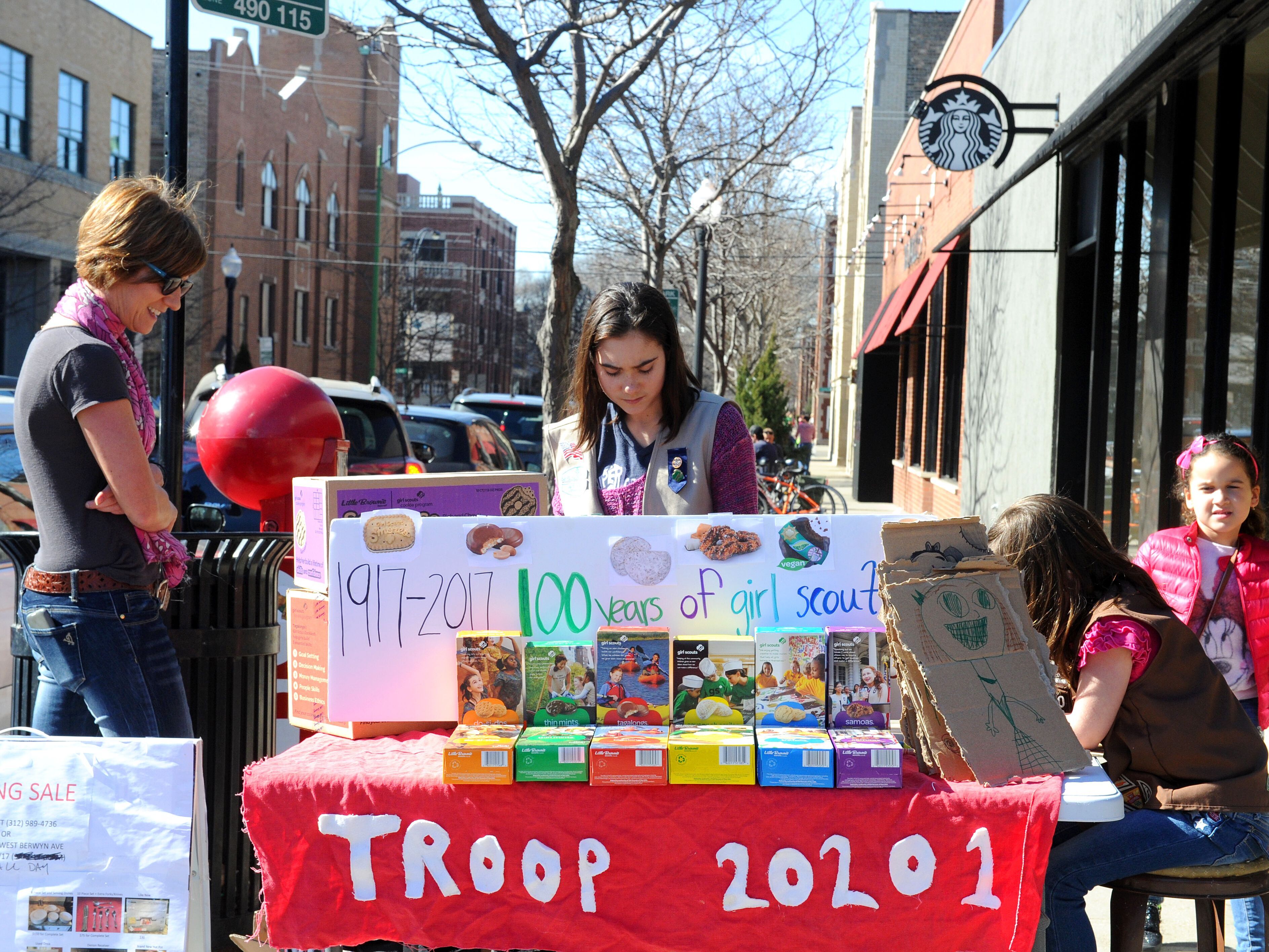 caption: Trina Sheridan, left, watches as her her daughters Molly and Edie sell cookies in Chicago in 2017, the 100th year of Girl Scout cookies.