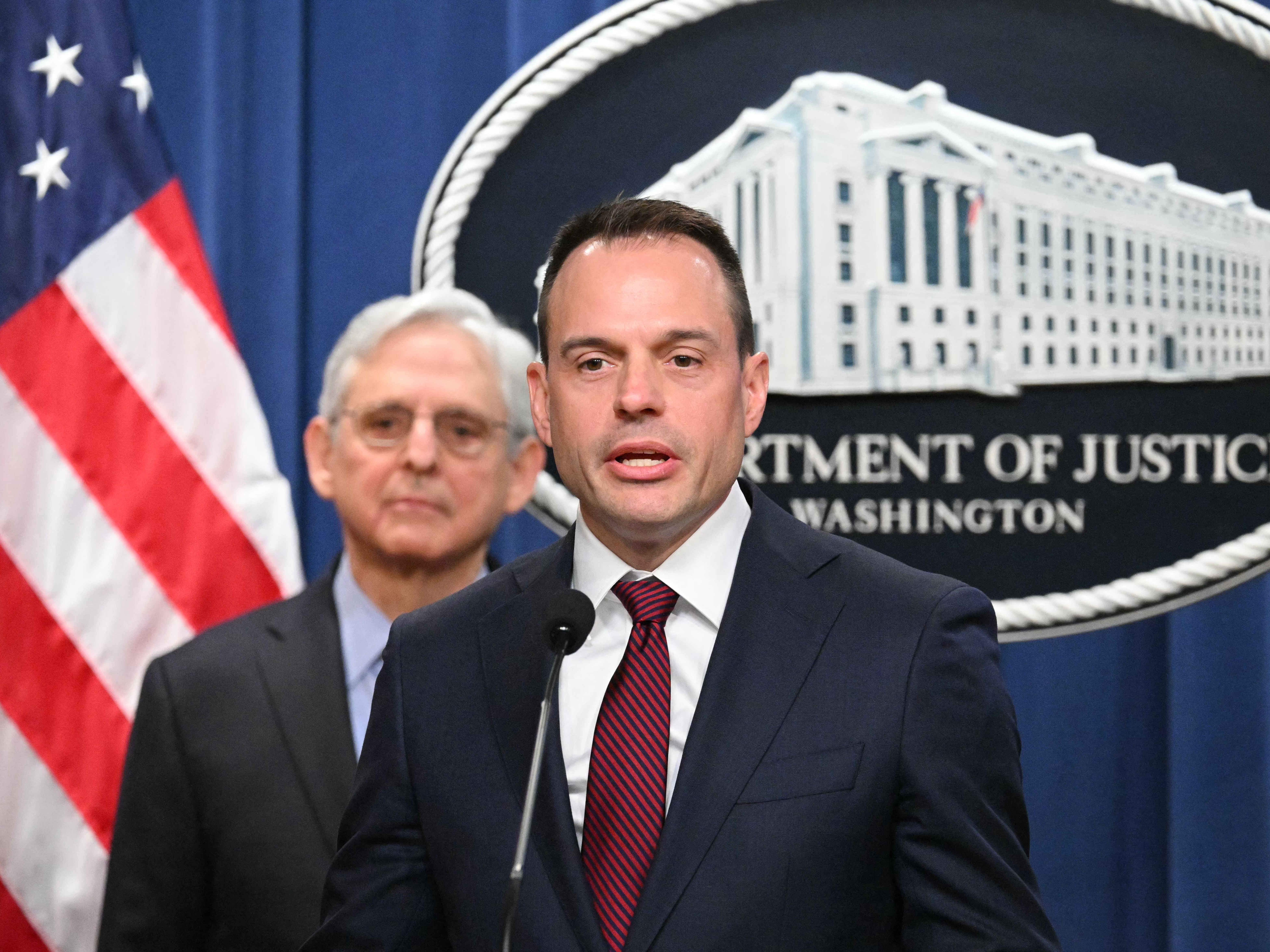 caption: Benjamin Mizer, with Attorney General Merrick Garland (left), speaks during a press conference announcing an antitrust lawsuit against Apple in March 2024.