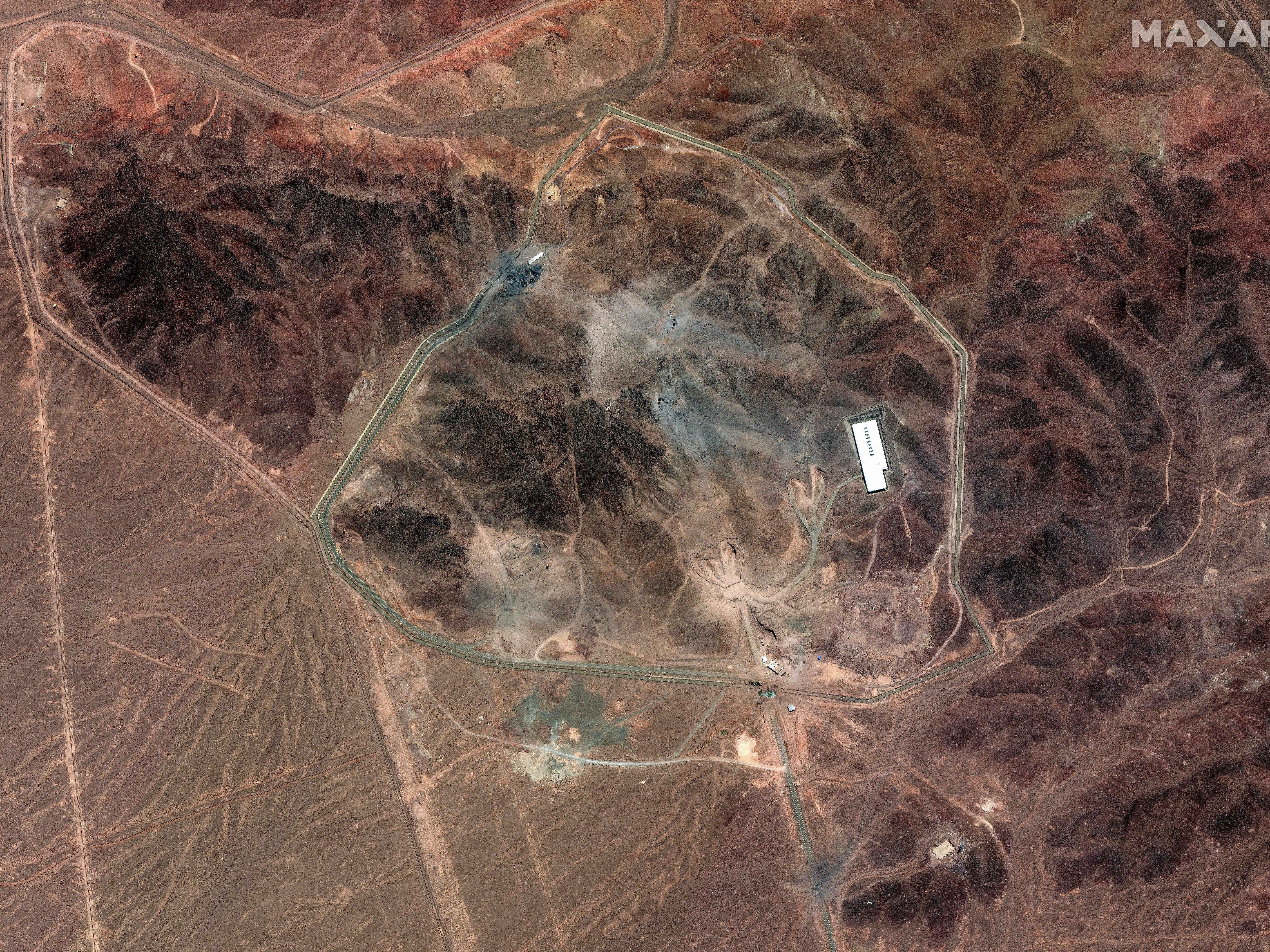caption: A satellite photo of Iran's Fordo fuel enrichment facility taken on June 24 shows debris (grey) from a U.S. strike employing several bunker-busting weapons. The Israeli Air Force destroyed additional roads and surface facilities in a subsequent strike.