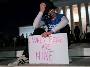 caption: A woman, mourning the death of Ruth Bader Ginsburg, holds a sign at the Supreme Court that reads, "when there are nine," something Ginsburg said to describe when there'd be enough women on the court.