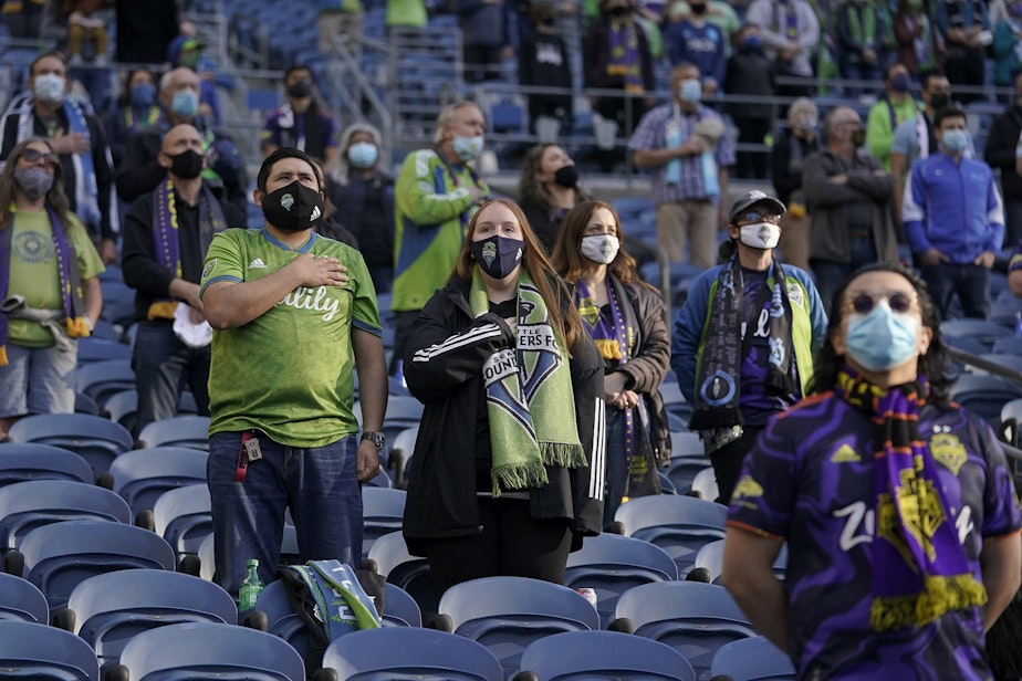 caption: Seattle Sounders fans wear masks as they stand during the singing of the national anthem before an MLS soccer match against the Los Angeles Galaxy, Sunday, May 2, 2021, in Seattle. 