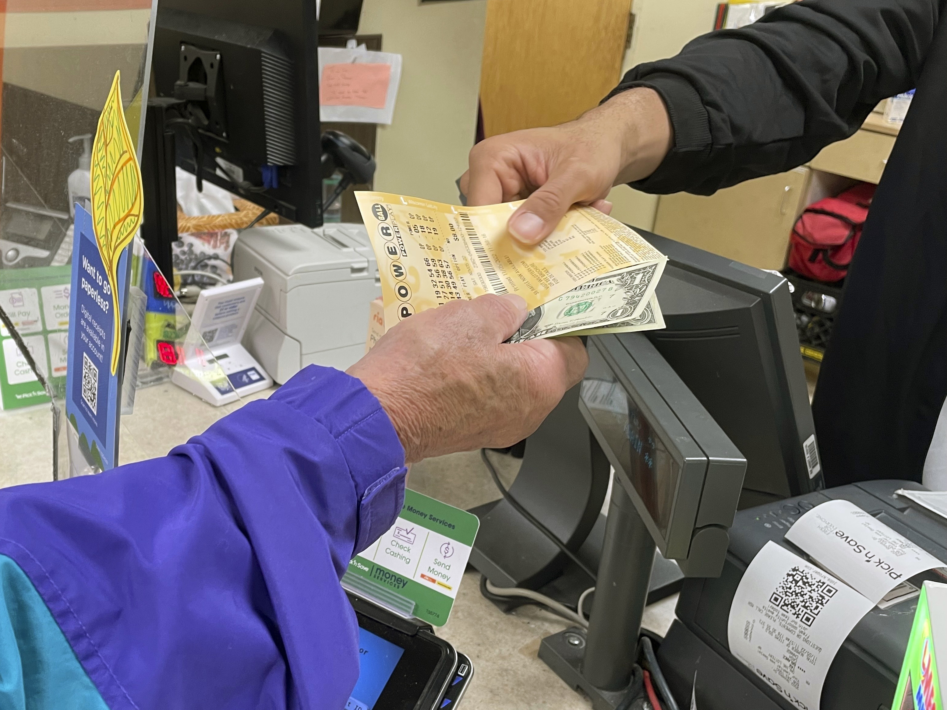 caption: From behind the lottery counter at a Pick 'n Save store in Madison, Wis., Djuan Davis hands Powerball tickets to Arpad Jakab, a retired utility worker who said it's his first time buying them.