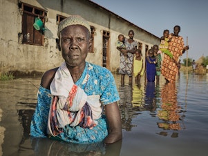 caption: Nyayua Thang, 62, left, stands waist-deep in the floodwaters in front of an abandoned primary school in South Sudan. Members of her village, displaced by extreme flooding as a result of heavy rainfall, are using the building as a refuge. Only small mud dikes at the entrance of the door are keeping the water out. (November 2020)
