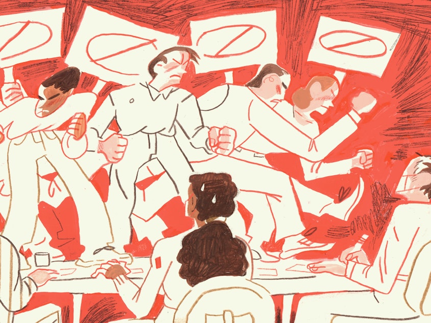 People stand on a table, protesting angrily, while others sit in chairs, frightened.