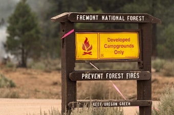 caption: A fire warning sign is seen amid trees which smolder and burn in Division Echo Echo of the Bootleg Fire on July 25, 2021, in the Fremont National Forest of Oregon.