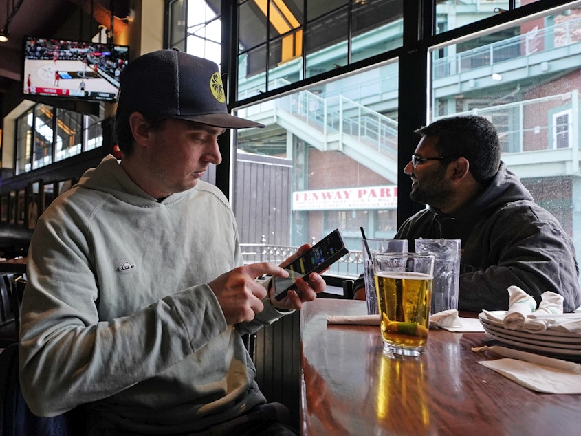 caption: Taylor Foehl (left), of Boston, looks at a mobile betting app on his phone after placing a wager, while watching a men's college basketball game at the Cask 'N Flagon sports bar on March 10, 2023, near Fenway Park in Boston.