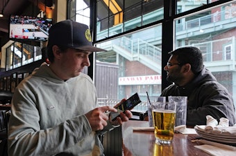 caption: Taylor Foehl (left), of Boston, looks at a mobile betting app on his phone after placing a wager, while watching a men's college basketball game at the Cask 'N Flagon sports bar on March 10, 2023, near Fenway Park in Boston.