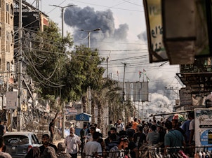 caption: People walk along a street as a plume of smoke rises in the background during an Israeli strike on the Bureij refugee camp in the Gaza Strip on Thursday, as battles between Israel and the Palestinian Hamas movement continue.