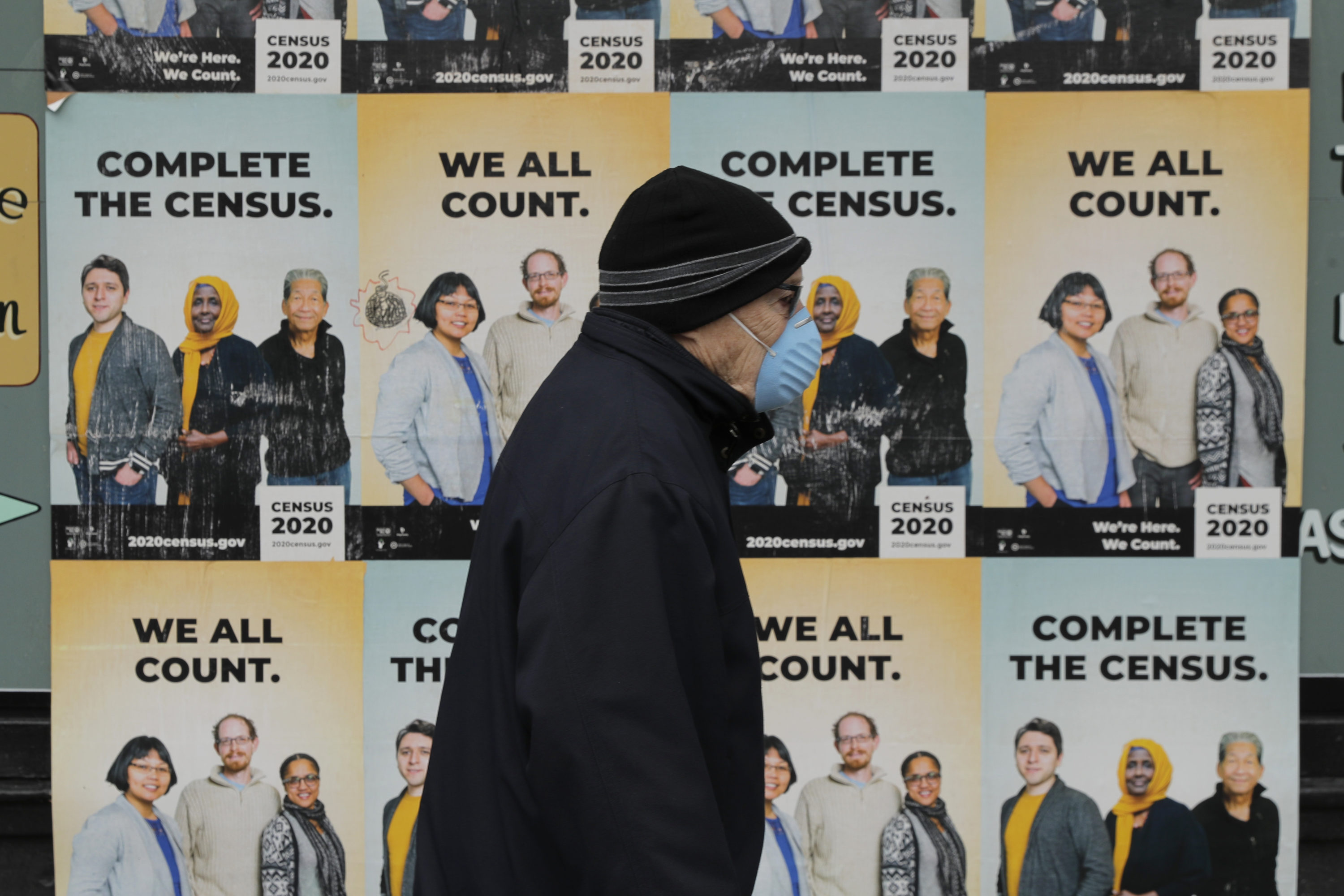caption: A man wearing a mask walks past posters encouraging participation in the 2020 Census, Wednesday, April 1, 2020, in Seattle's Capitol Hill neighborhood. (Ted S. Warren/AP)