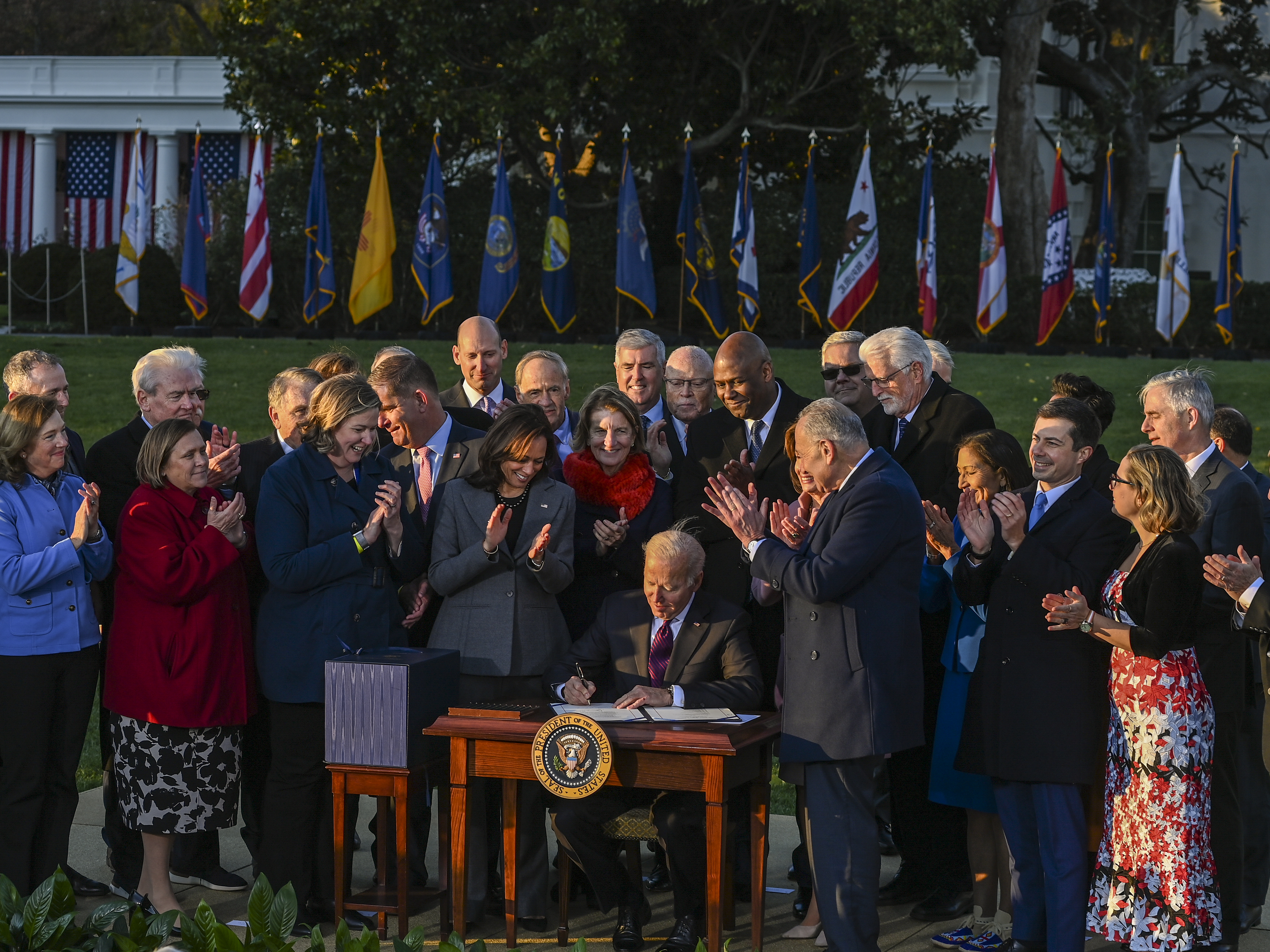 caption: President Biden signs the Infrastructure Investment and Jobs Act as he is surrounded by lawmakers and members of his Cabinet during a ceremony on the South Lawn at the White House on Monday.