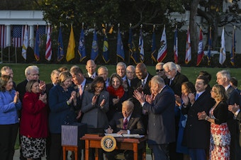 caption: President Biden signs the Infrastructure Investment and Jobs Act as he is surrounded by lawmakers and members of his Cabinet during a ceremony on the South Lawn at the White House on Monday.