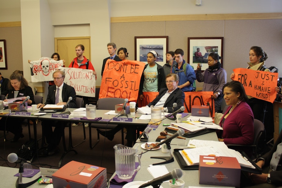 caption: Protesters hold signs around a table populated by UW Regents.