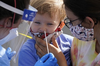 caption: Penny Brown, 2, is held by her mother, Heather Brown, as her nose is lightly swabbed during a test for COVID-19 at a new walk-up testing site at Chief Sealth High School, Friday, Aug. 28, 2020, in Seattle. The child's daycare facility requires testing for the virus. The coronavirus testing site is the fourth now open by the city and is free. 
