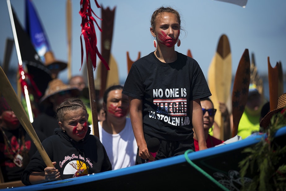 caption: Rosalie Fish, 18, requests permission to come ashore in a Cowlitz tribal canoe after a multi-day canoe journey on Wednesday, July 24, 2019, during Paddle to Lummi at the Lummi Nation Stommish Grounds. A red painted hand print over her mouth honors missing and murdered indigenous women. "The purpose of the paint here today is to show a symbol of unity and to say that my canoe family stands with the relatives that have been affected by this epidemic," Fish said. 