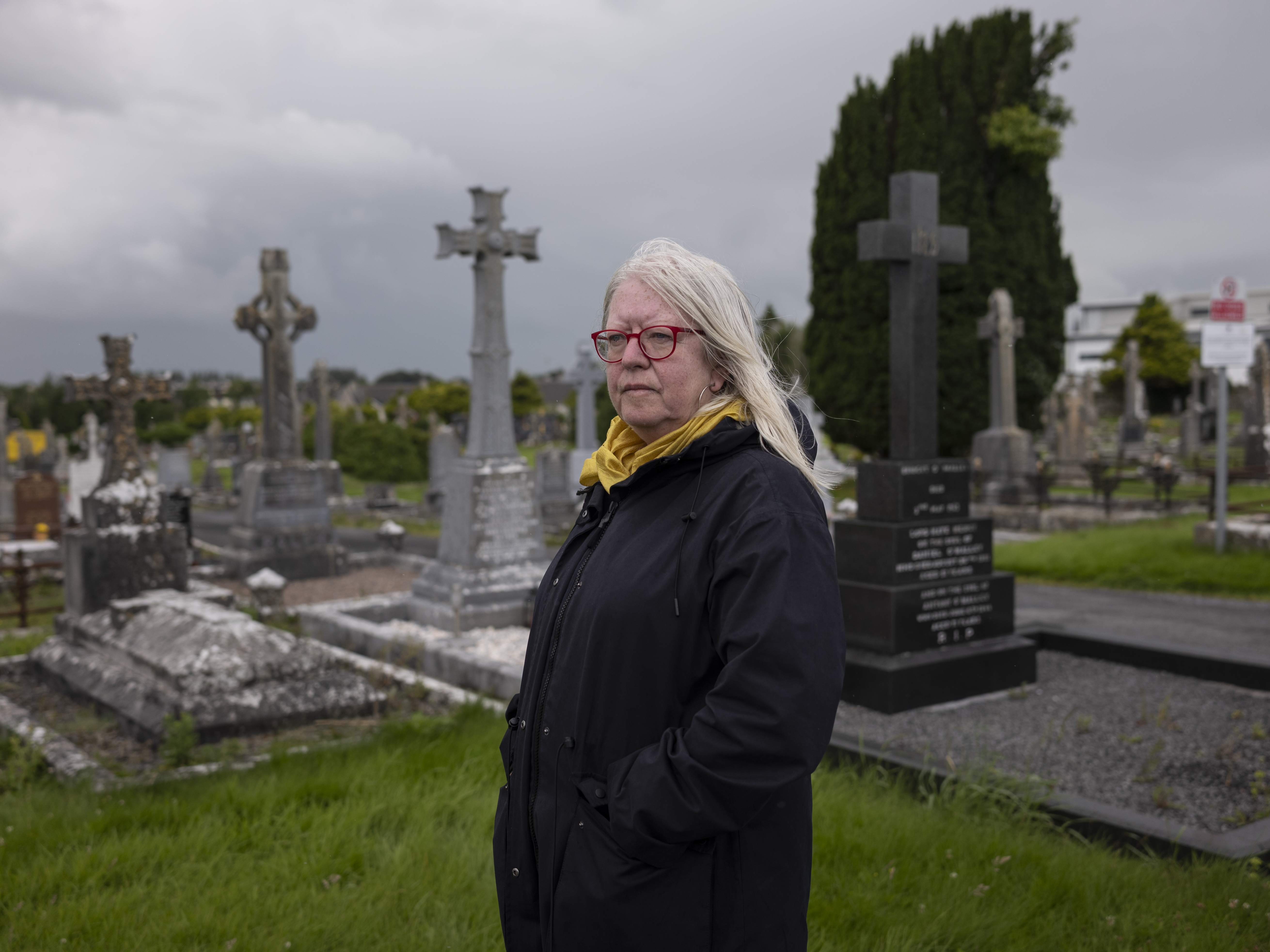 caption: Anna Corrigan stands in the Tuam graveyard, near the site of a former maternity institution for unmarried mothers and their children run by the Bon Secours religious order in Tuam, Ireland, on July 14. For years, Corrigan has sought answers about the fate of her two missing siblings, born at the institution.