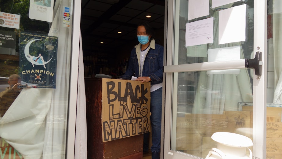 caption: Suthap Manivong sells wine and offers recommendations from behind a makeshift counter in the doorway of Champion Wine Cellars in Greenwood. Like many businesses across the city, Champion Wine Cellars chose to add a handmade sign displaying support for the Black Lives Matter movement to the pandemic-related notices taped the the front door. Manivong is the co-owner of the 51-year-old wine store: the oldest in Seattle.