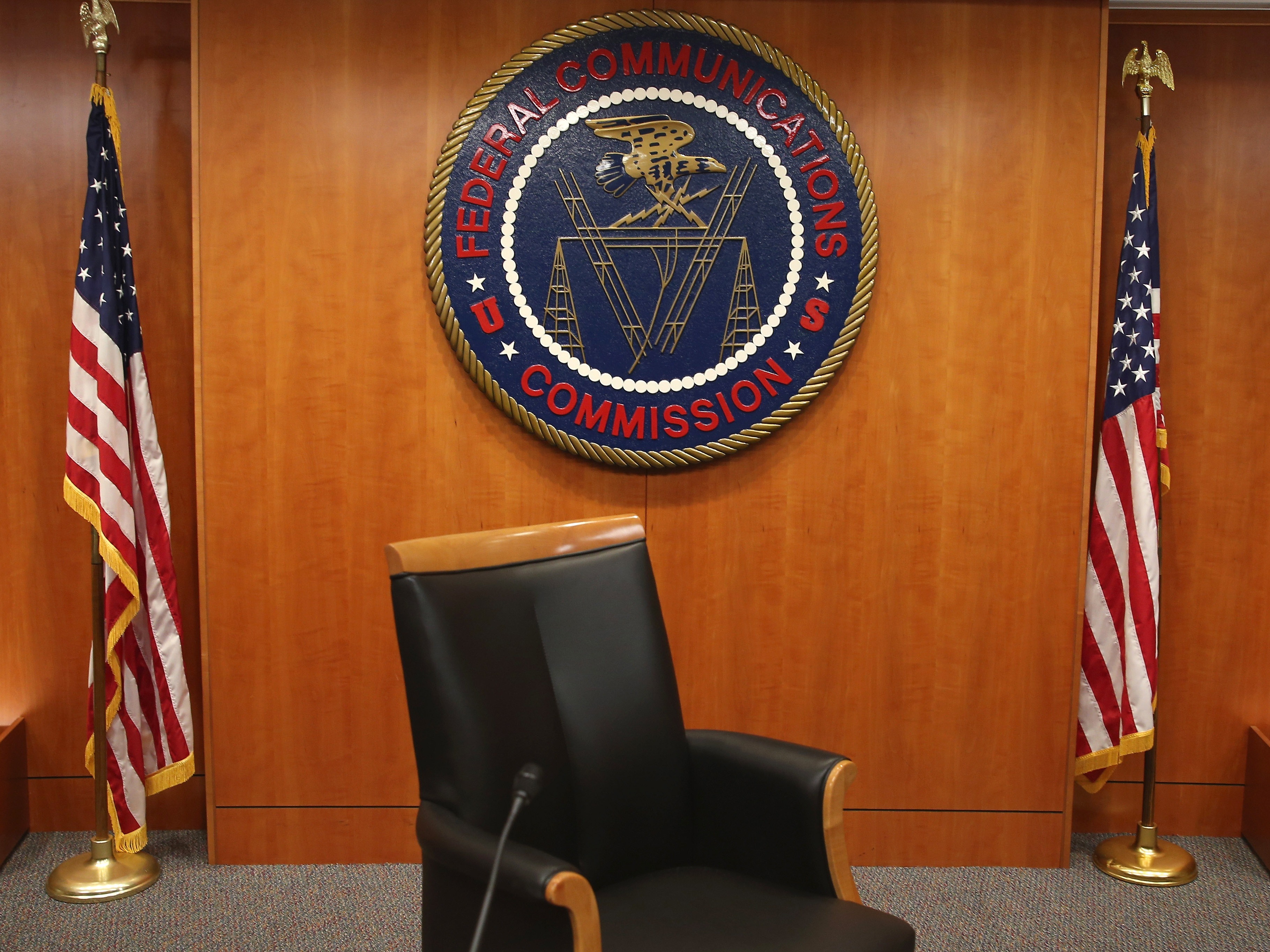 caption: The seal of the Federal Communications Commission hangs between two American flags; the FCC is urging broadcasters to air more "patriotic" content in the run-up to the country's 250th celebrations this summer.