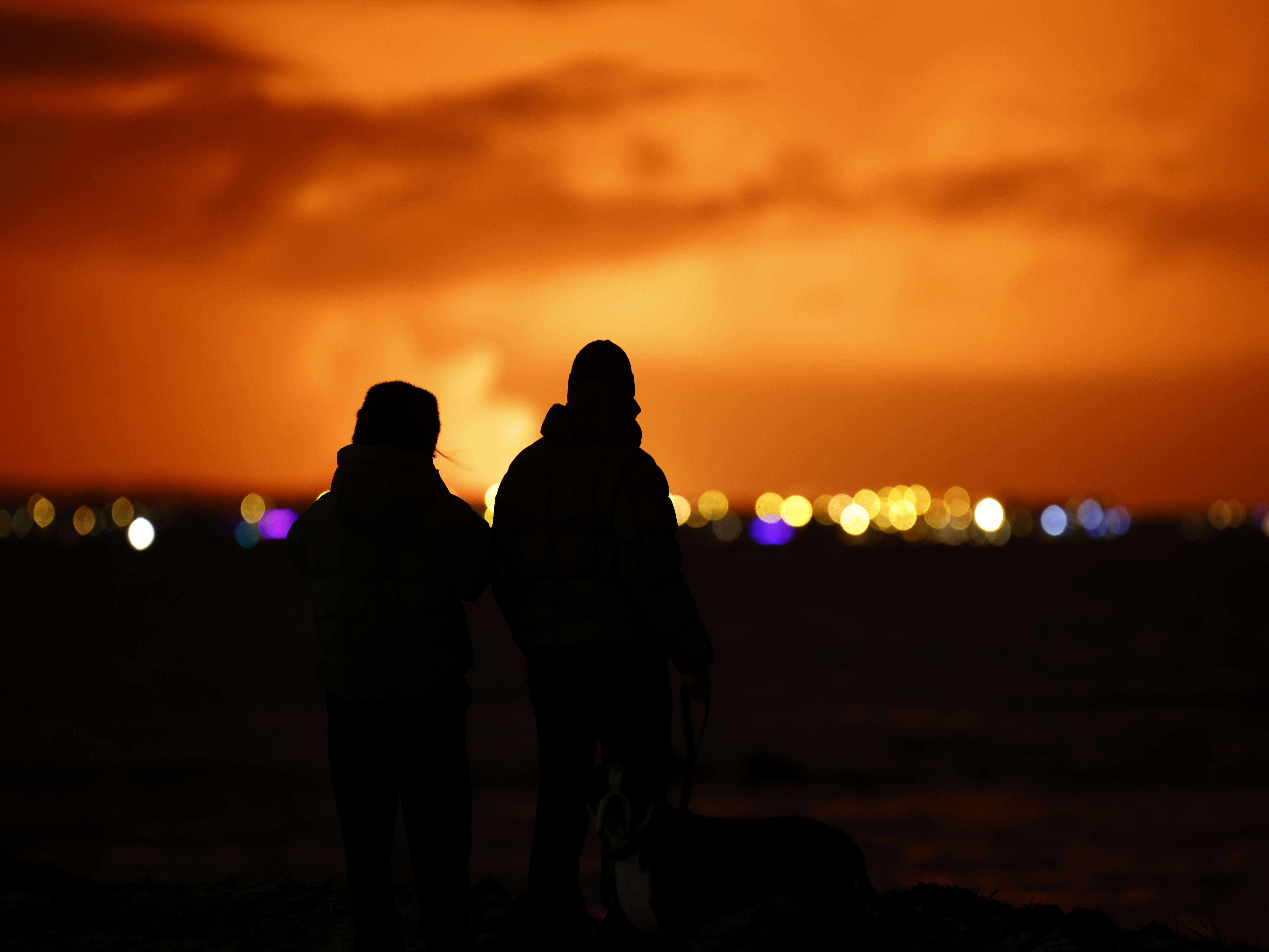 caption: People watch as the night sky is illuminated caused by the eruption of a volcano on the Reykjanes Peninsula in southwestern Iceland seen from the capital city of Reykjavik on Monday, Dec. 18, 2023.