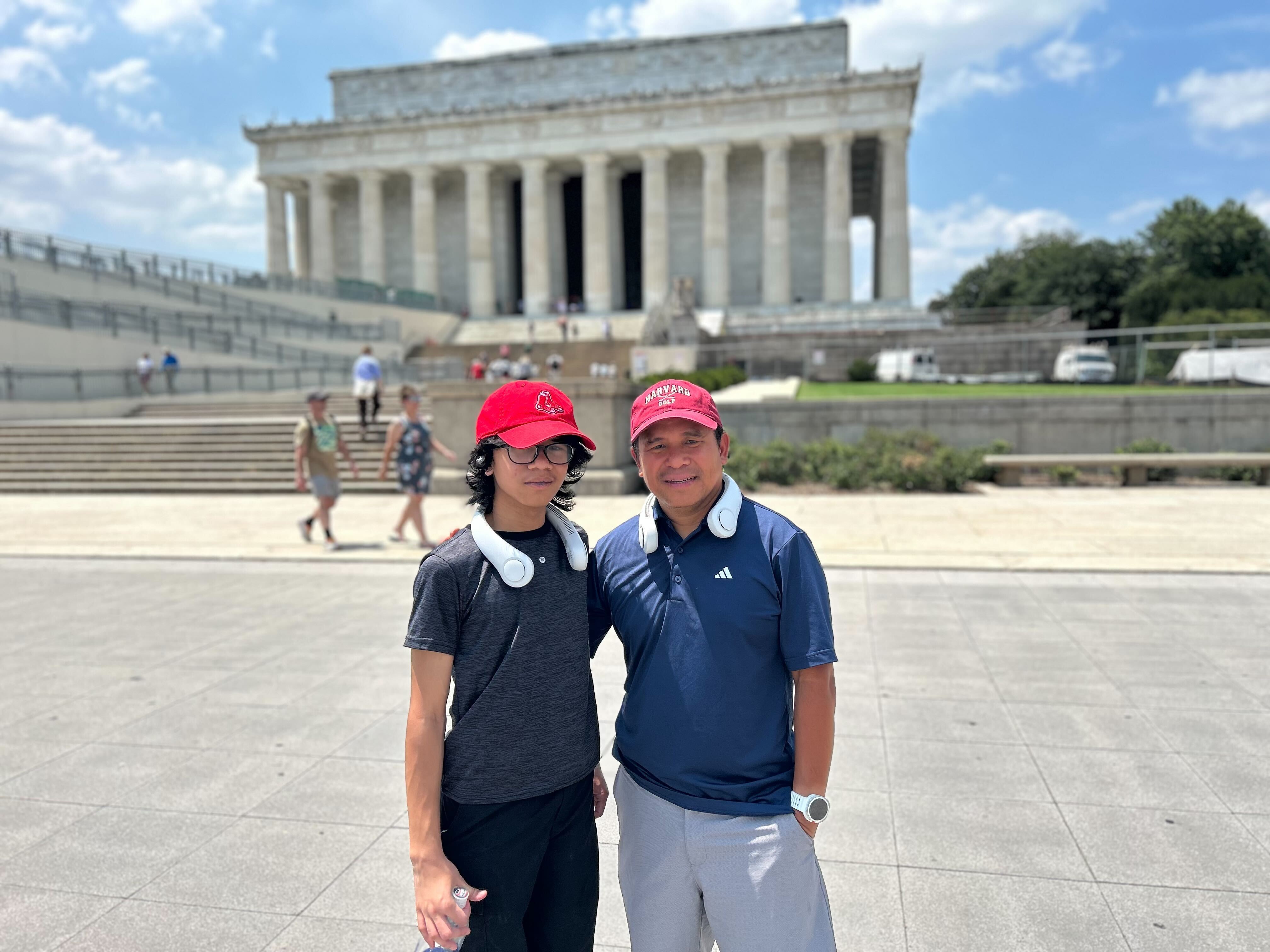 caption: Jeffrey Pagulong and his son Christof stand in front of the Lincoln Memorial in Washington, D.C., on July 29. Pagulong, who is visiting from Boston, said his neck fan "helps a lot" in the high temperature. It makes noise, he said, but the noise is better than the heat.