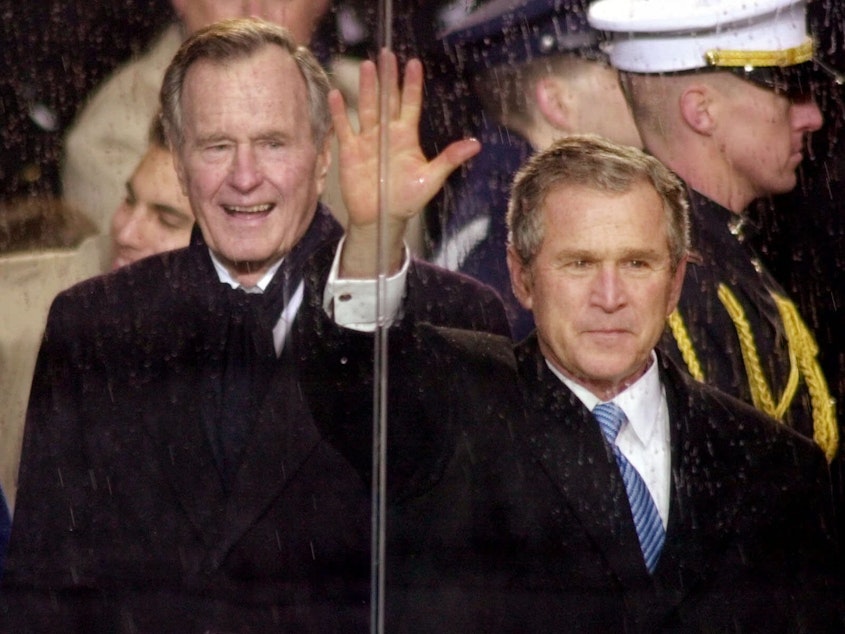 caption: In this Jan. 20, 2001, file photo, standing in the rain, President George W. Bush waves as he watches his inaugural parade pass by the White House viewing stand in Washington, Saturday afternoon, Jan. 20, 2001. With him are his wife and first lady Laura Bush and his father, former President George H.W. Bush.