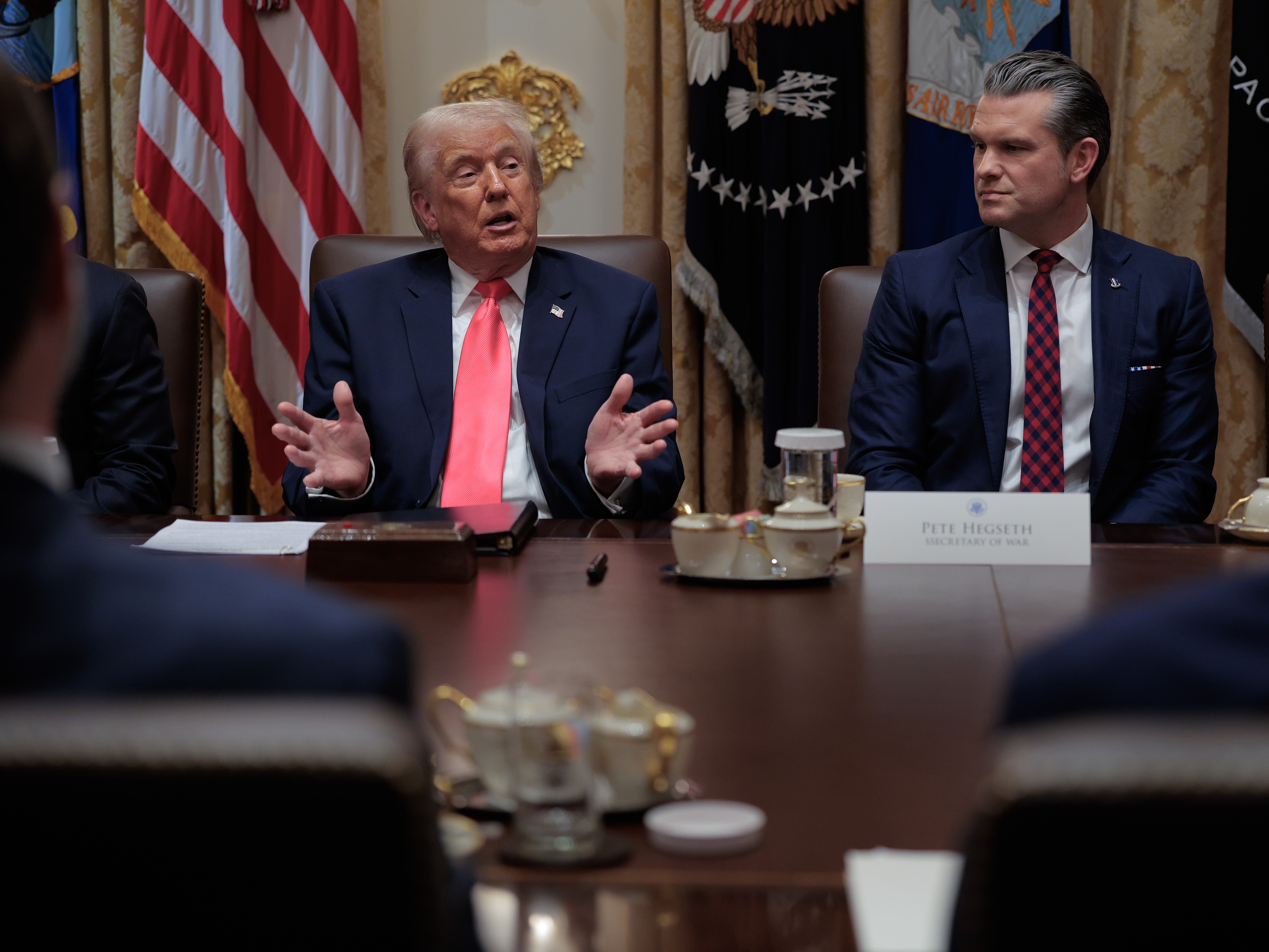 caption: President Trump speaks as U.S. Secretary of Defense Pete Hegseth looks on during a meeting of his Cabinet at the White House in December 2025.