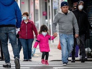 caption: People, some wearing masks, walk down a street in the Corona neighborhood of Queens in New York on Tuesday.