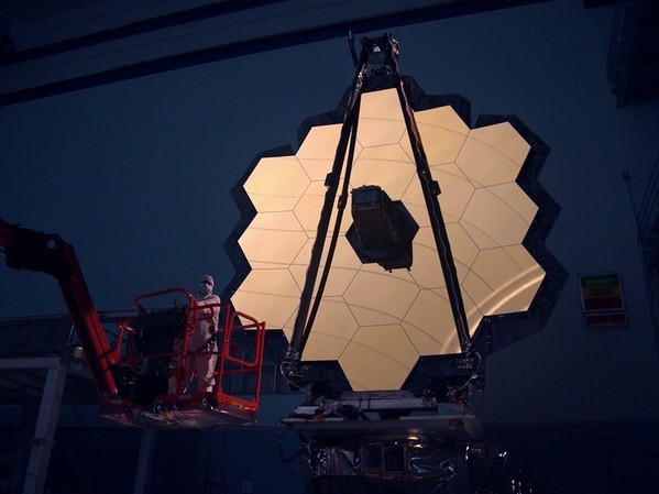 caption: The James Webb Space Telescope's primary mirror is illuminated inside a darkened clean room. The entire telescope is now packed inside a rocket, awaiting launch.
