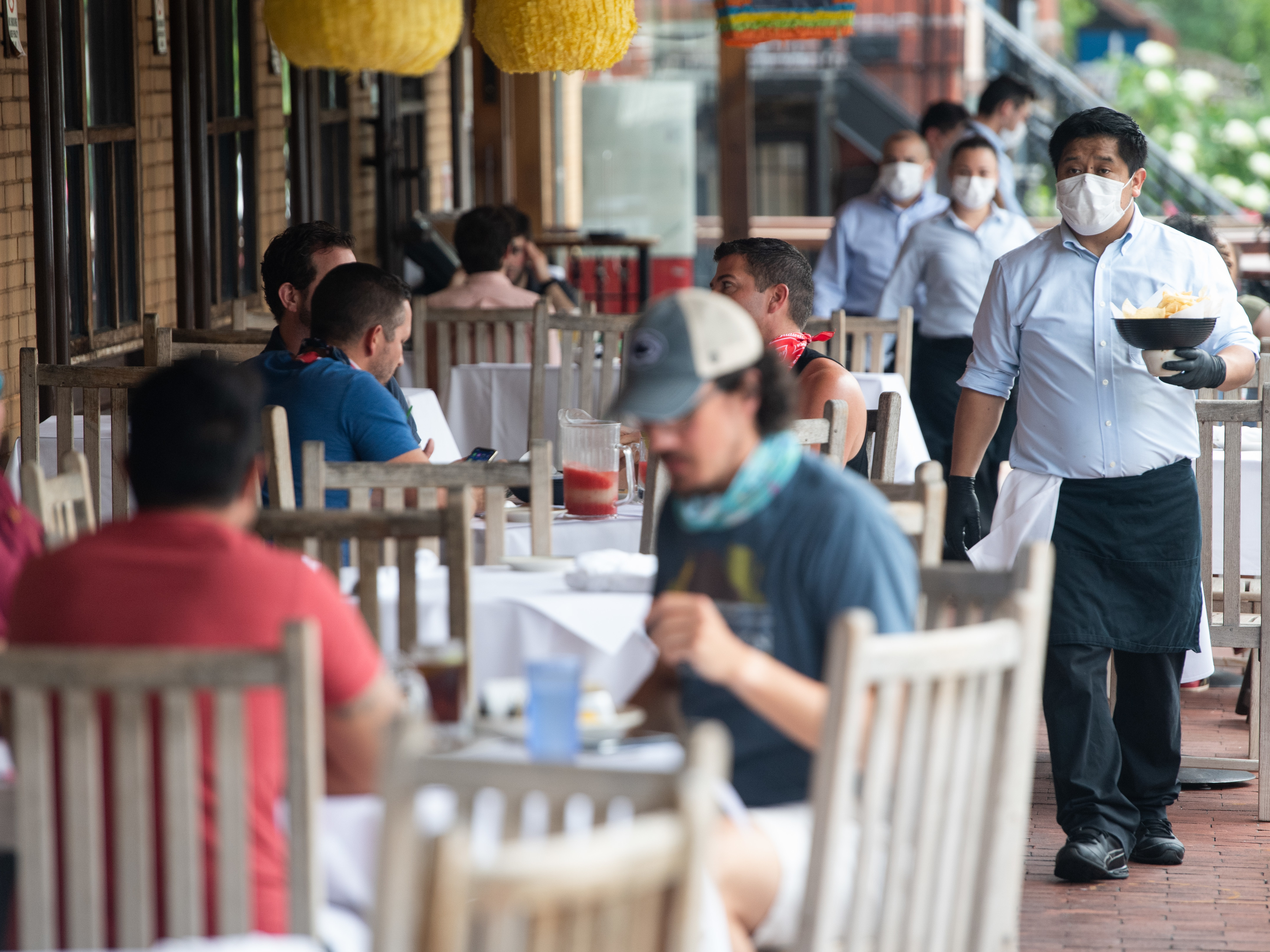 caption: A waiter wearing a mask and gloves delivers food to a table to customers seated at an outdoor patio at a restaurant in Washington, D.C. in May.