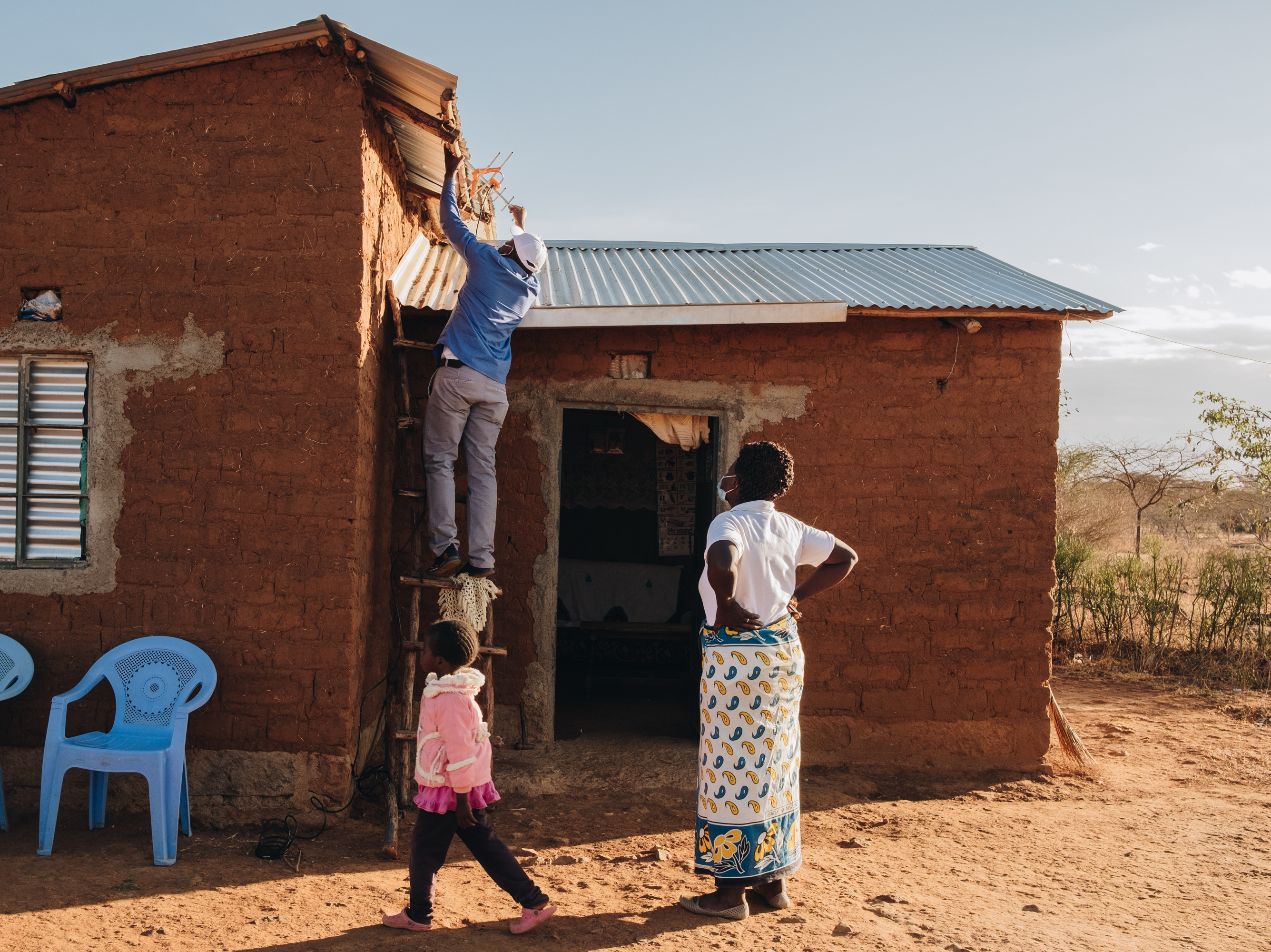 caption: Winifred Muisyo, right, and her five-year-old daughter, Patience Kativa, watch Stanlas Kisilu, left, as he installs a TV tuner on the roof of her home. The TV is connected to a solar panel provided by d.light, a company partially funded by climate financing from wealthier nations.