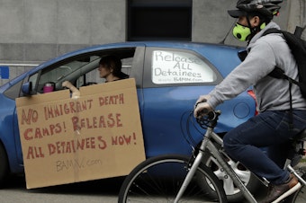 caption: A masked cyclist passes a motorist protesting in a vehicle at a U.S. Immigration and Customs Enforcement field office last month in San Francisco.