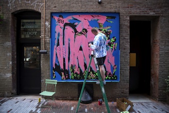 caption: Devin Liston works on a mural titled ‘Dogpile’ outside of the RailSpur buildings ahead of MLB All-Star week on Thursday, July 6, 2023, in Seattle’s Pioneer Square neighborhood. 