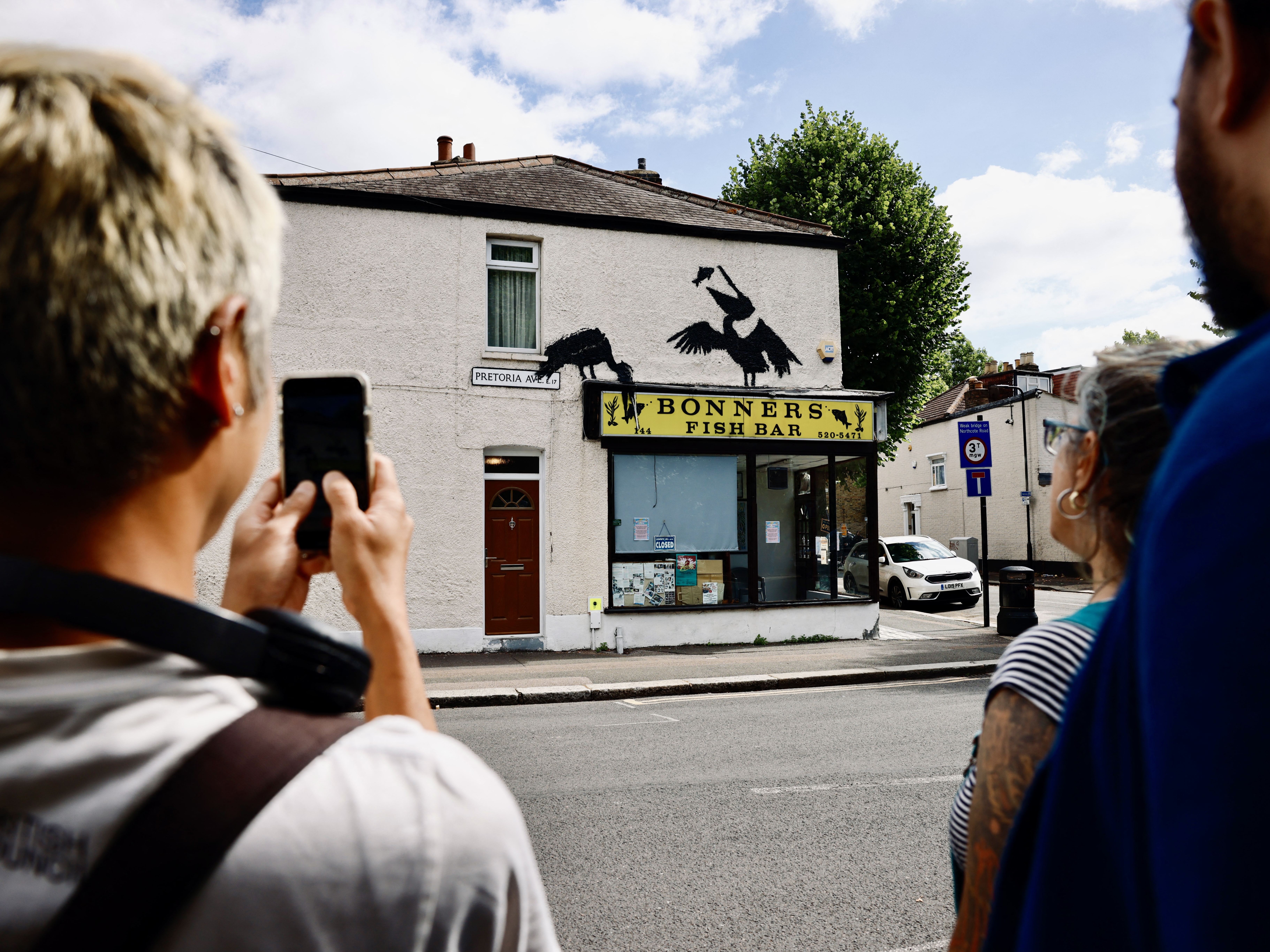 caption: People gather to look at an artwork by street artist Banksy depicting two pelicans catching fish, painted on top of a fish-and-chips shop in Walthamstow, northeast London, on Aug. 9.